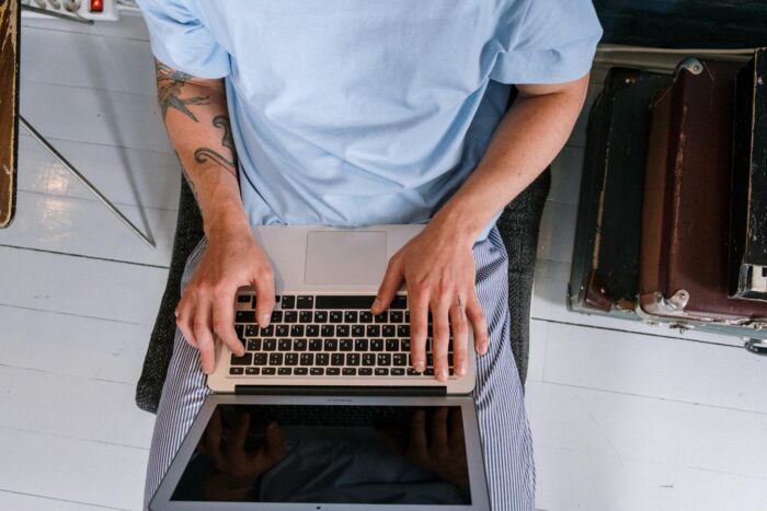 An adult with tattoos typing on a laptop indoors, viewed from above.